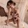 Woman wearing forbidden elegance lace -inspired bodysuit with floral print, kneeling on bed in romantic bedroom setting.