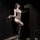 Woman wearing golden night seductive lace bodysuit with sheer mesh, holding a whip in dimly lit room.