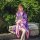 A woman wearing a purple lolita dress with cherry blossom patterns, paired with a pink ribbon and fan, sitting by a pond surr...