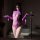 Woman in purple sheer bodysuit with lace mesh, long sleeves, and mask, posing seductively in bathroom setting.