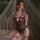 Woman wearing mannyan black lace bodysuit with bow detail and garters, posing on bed with sheer canopy background.