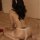 Woman wearing mannye qing feathered backless thong bodysuit with bow detail, seated on bed in seductive wife style outfit.
