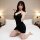 A woman poses on a bed wearing a black midnight allure deep v-neck slip dress, featuring a seductive wine red satin finish wi...