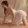 A woman poses in a sheer lace nightgown with backless straps, paired with matching delicate lingerie, set against soft draper...