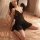 A woman poses on a bed wearing a black sheer lace nightgown with backless straps, paired with matching lingerie, creating an ...