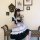 A person wearing a black and white lolita maid dress with ruffles, sitting on a bench holding flowers against a vintage wall ...