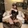 Woman wearing black vinyl and lace open-crotch bodysuit with thigh-high stockings, posing on round ottoman in bedroom setting...