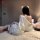 A woman sits on a bed wearing a luxurious white silk nightgown with lace detailing, paired with matching sheer mesh sleepwear...