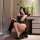 A woman poses elegantly in a black lace cami dress featuring a side slit design, holding a drink while seated indoors near fl...