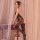 Woman wearing black lace mesh bodystocking with open crotch, sheer stockings, and seductive design in soft bedroom setting.