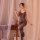 Woman modeling black sheer mesh bodystocking with lace straps and open crotch, posing by bedside table in soft lighting.