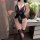 Woman in black lace bodysuit with sheer mesh and open-crotch design, holding wine glass beside floral vase.