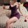 Woman in purple lace nightsiyese sleep dress with sheer mesh, holding wine glass on fluffy rug with rose petals.