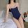 A woman poses on a bed wearing a navy blue one-piece swimsuit with white trim, featuring thin straps and a sleek design, perf...