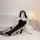 Model wearing black plus size punishment nun lingerie set with cut-outs and high slit, posing on bed for sexy role play.