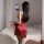 Woman in red satin chemise nightgown with adjustable straps, posing by bed, holding phone for mirror selfie.