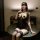 Woman in black sheer lingerie set with lace details, wearing a mask and sitting on a round table.