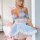 Woman wearing light blue pure & playful lingerie sleep dress with ruffles, bow details, and waist cinching in a cozy bedroom.