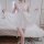 Woman wearing white sheer lingerie sleep dress with lace details, standing by bed in soft lighting.