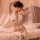 Woman wearing white lace babydoll nightgown with sheer mesh, adjustable straps, and bow detail, seated on bed with soft light...