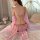 Woman wearing pink pure & seductive lingerie set with sheer sleeves and gold chain detail, seated on bed for couples’ rolepla...