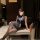 Woman in pure & seductive secretary style bodycon outfit, posing elegantly on a cushioned bench with mask and lanyard.