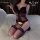 Woman modeling a black and purple lace lingerie set with an open-crotch vest bra and matching thigh-high stockings on a bed.