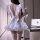 A woman wears a white cosplay maid costume with a bow backless design, pleated skirt, and thigh-high stockings, holding a ted...