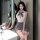 A woman poses in a bathroom wearing a white top with a gray bow tie, paired with a matching skirt and stockings, showcasing a...