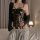 Woman wearing black lace lingerie set with sheer cups and bow detail, paired with gray sweatpants and holding a glass.