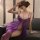 Woman wearing purple lace sleep dress with sheer mesh cami nightgown, sitting on couch in bedroom lingerie.