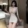 A woman poses elegantly in a white bodycon dress featuring a deep v-neckline and lace detailing, holding a glass of champagne...