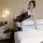 Woman wearing qingyegé black lace mesh bodysuit and sheer stockings, posing seductively on a bed in a modern bedroom.