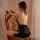 Woman wearing black backless sleep dress with lace details, seated on vintage chair in soft lighting.