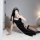 Woman in black seductive nun costume lingerie with lace trim, posing on white rug while taking mirror selfie.