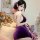 Woman wearing seductive purple cheongsam lingerie with open back, holding fan in cozy bedroom setting.