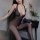 Woman wearing sheer black bodystocking with lace trim and open-crotch design, seated on chair beside small table with vase of...