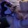 Woman in black strappy lingerie set with fishnet stockings, lying on bed under purple lighting.