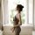 Woman wearing sensual sheer mesh bodysuit with backless neck-hanging design, standing by window in transparent sleepwear.