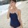 A woman stands on a beach wearing a navy blue one-piece swimsuit with white trim, featuring a halter neck design and thin str...