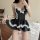 Woman wearing black lace-trimmed maid-style sleep dress with white bows and ruffles, posing on bed.
