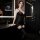 Woman wearing black lace bodysuit with sheer mesh, stockings, and skirt – seductive lingerie set in modern bathroom.