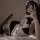 Woman in black and white maid costume with halter neck bow, lace trim, and satin apron, lying on bed taking mirror selfie.