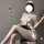 Woman in white nurse costume with bow-tie skirt and round neck top, holding tablet, seated on stool.