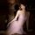 Woman wearing pink sheer lace sleep dress with underwire support and chain detail, posing on bed in dim lighting.