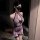 Woman wearing a purple lace cheongsam lingerie set with sheer panels, bow detail, and fishnet stockings in a dimly lit room.