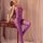 Woman in shiny purple mesh bodystocking with open-crotch design, leaning on wooden desk beside books and sheer curtains.
