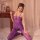 Woman wearing purple sheer bodystocking with open-crotch design, kneeling on bed beside wooden nightstand with books and lant...