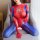 Woman in red and blue spider-themed sexy bodysuit with open-crotch design, sitting on a gray chair.