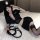 Woman in black police-themed lingerie set with short skirt, cap, and fishnet stockings, holding a whip on a bed.